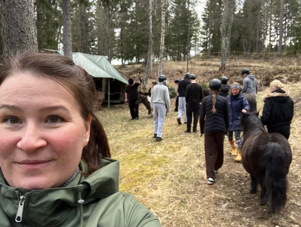 Opettaja katsoo kameraan hymyillen. Takana nuoria kävelemässä ponien kanssa metsäisessä ympäristössä.